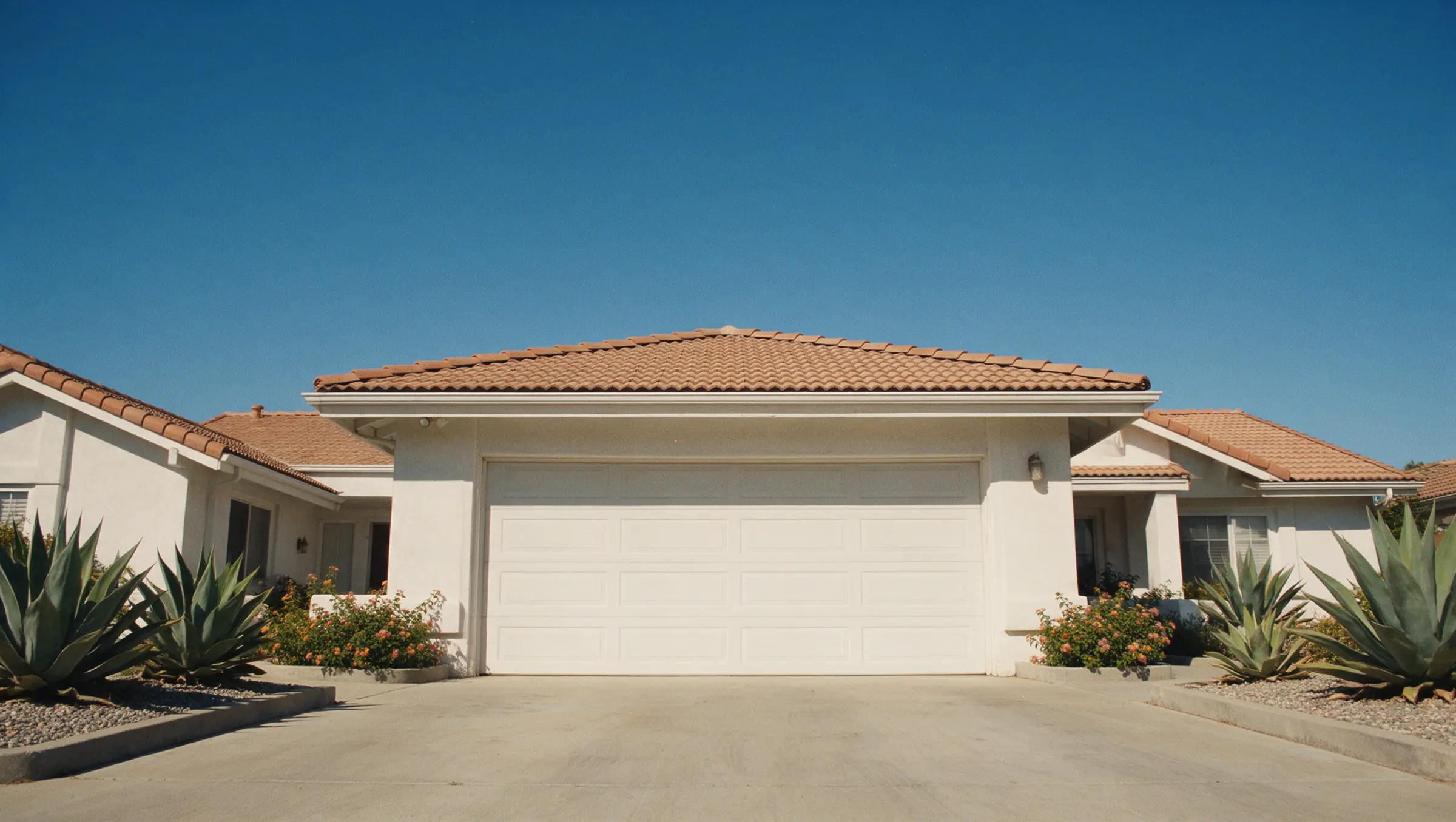 A white stucco ranch house in Corona with a closed two-car garage door, hard afternoon sun.